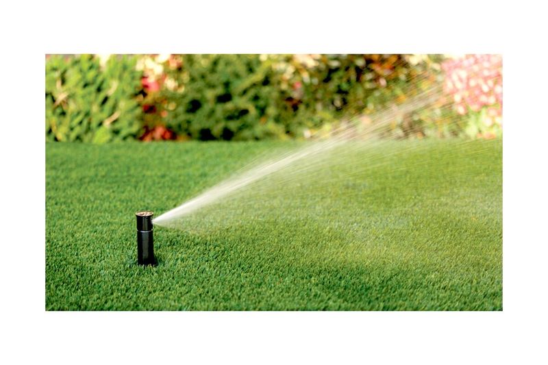 Aspersor para jardín ajustable 1/2 pulgada Rain Bird, instalado en césped verde emitiendo un chorro de agua en arco