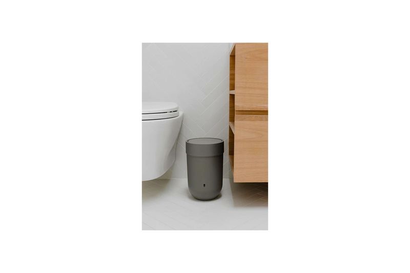 Papelera para baño gris moldeado Plast Umbra, ubicada en el suelo junto a un inodoro blanco y un mueble de madera