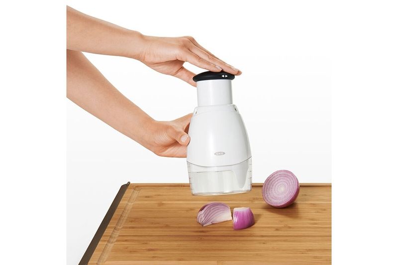 Picador de alimentos pequeño crema Oxo, siendo utilizado por una persona sobre una tabla de madera con trozos de cebolla