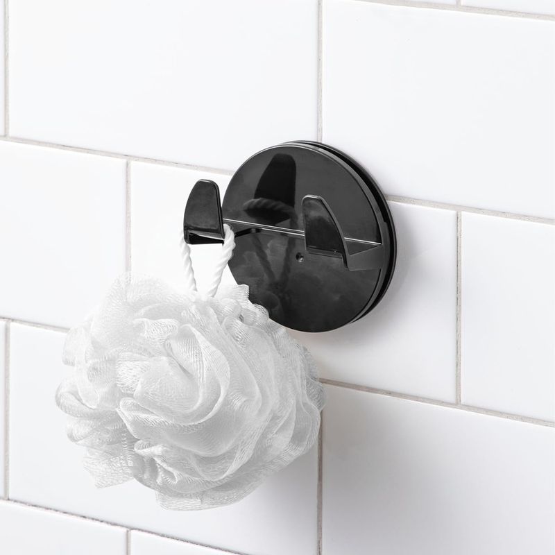 Gancho para baño autoadhesivo doble negro mate Interdesign, instalado en pared de azulejos blancos con una esponja blanca