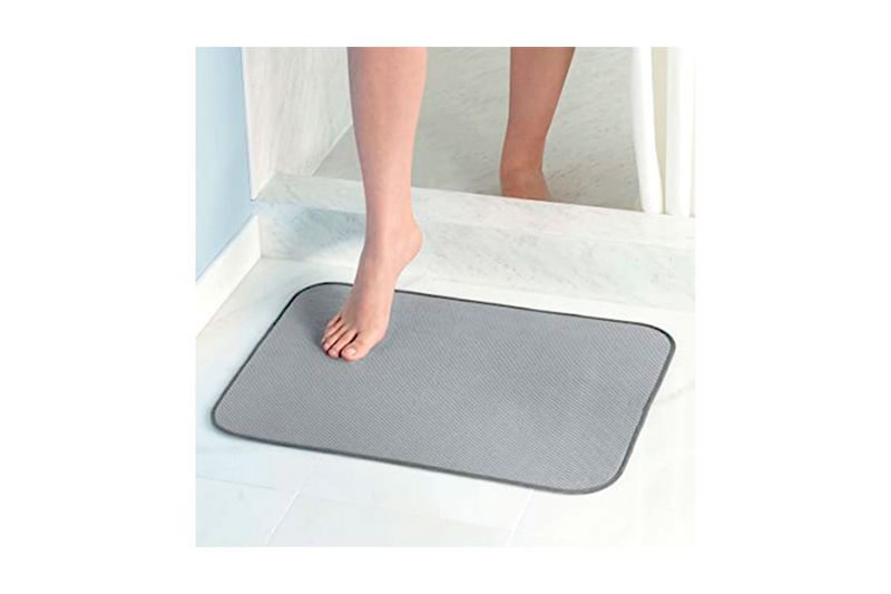 Alfombra de baño grey Idry microfibra 18 x 24 pulgadas Interdesign, colocada en el piso con un pie apoyado sobre ella