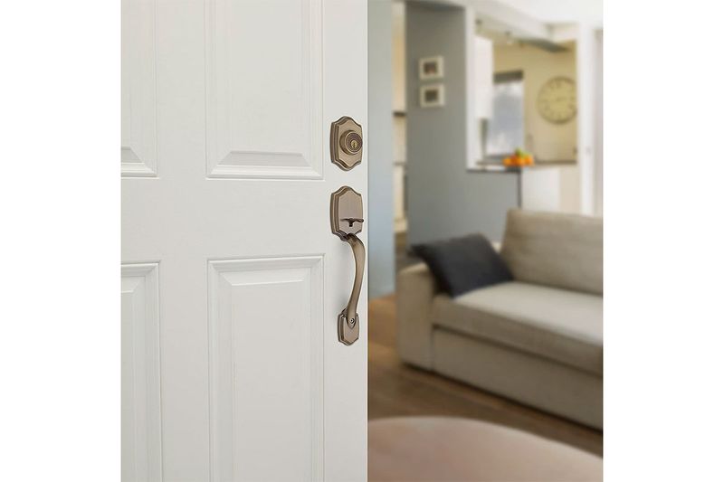 Cerradura pichel Belleview llave/mariposa bronce antiguo Kwikset, instalada en puerta blanca con fondo de sala desenfocado