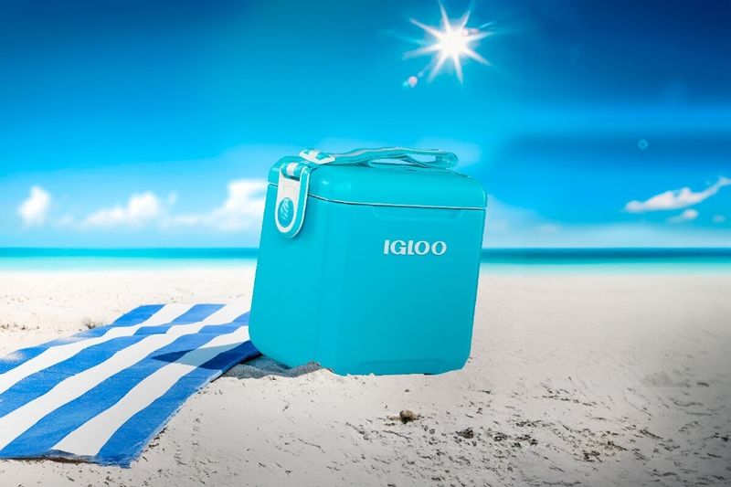 Hielera 11 qt turquesa Tag a Long con correa Igloo, situada sobre arena blanca de playa con una toalla azul al lado