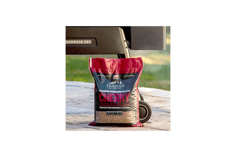 Pellets de madera cherry 20 lb Traeger, bolsa cerrada apoyada en el suelo junto a una parrilla Ironwood 885