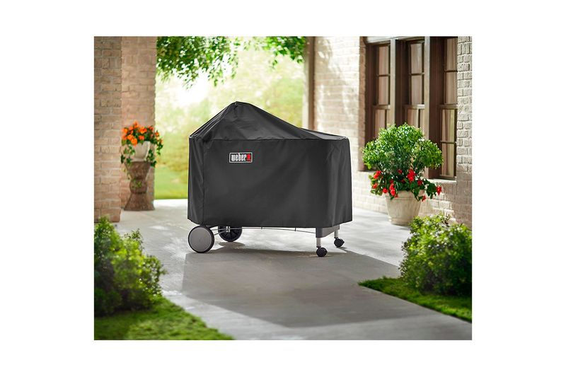Funda para barbacoa Premium/Deluxe carbón negra Weber, colocada sobre la barbacoa en un patio exterior con plantas