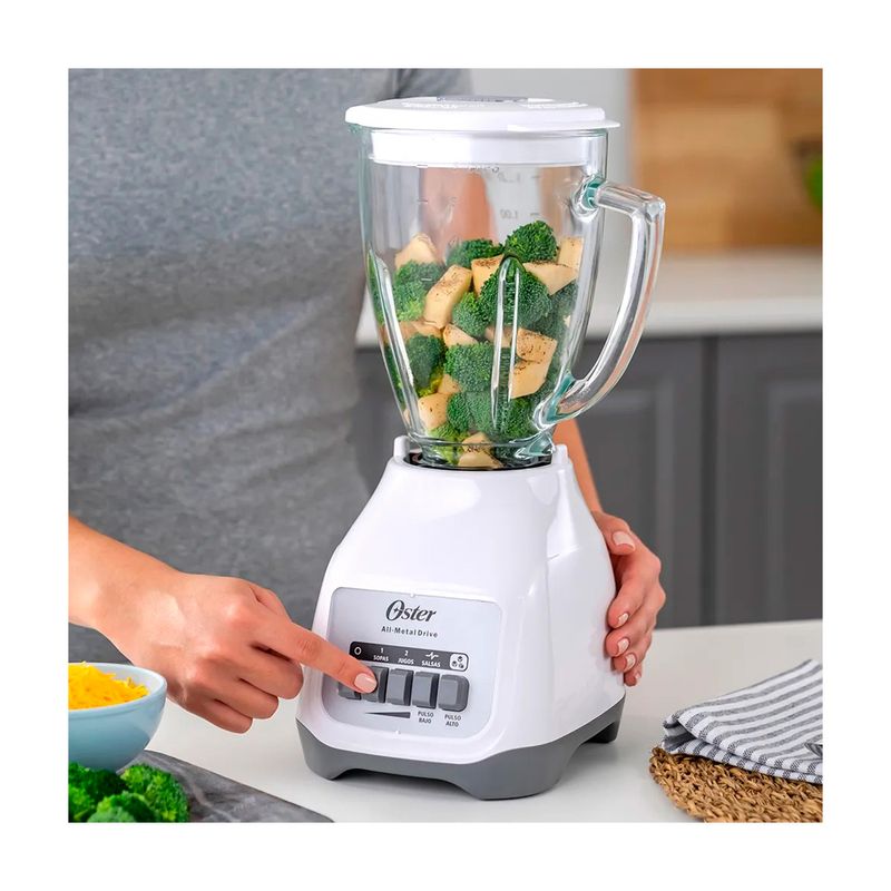 Licuadora vaso vidrio 1.5 l 800 W 2 vel blanca Oster, persona operando los botones de control con el vaso lleno de brócoli
