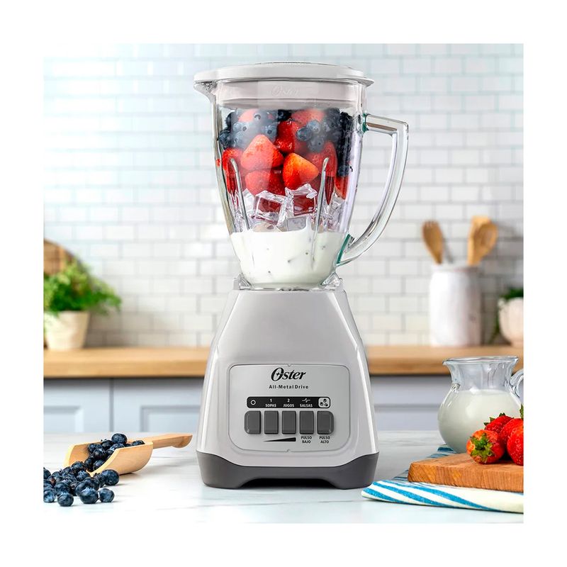 Licuadora vaso vidrio 1.5 L 800 W 2 vel blanca Oster, en uso con frutas y hielo en el vaso sobre una cocina