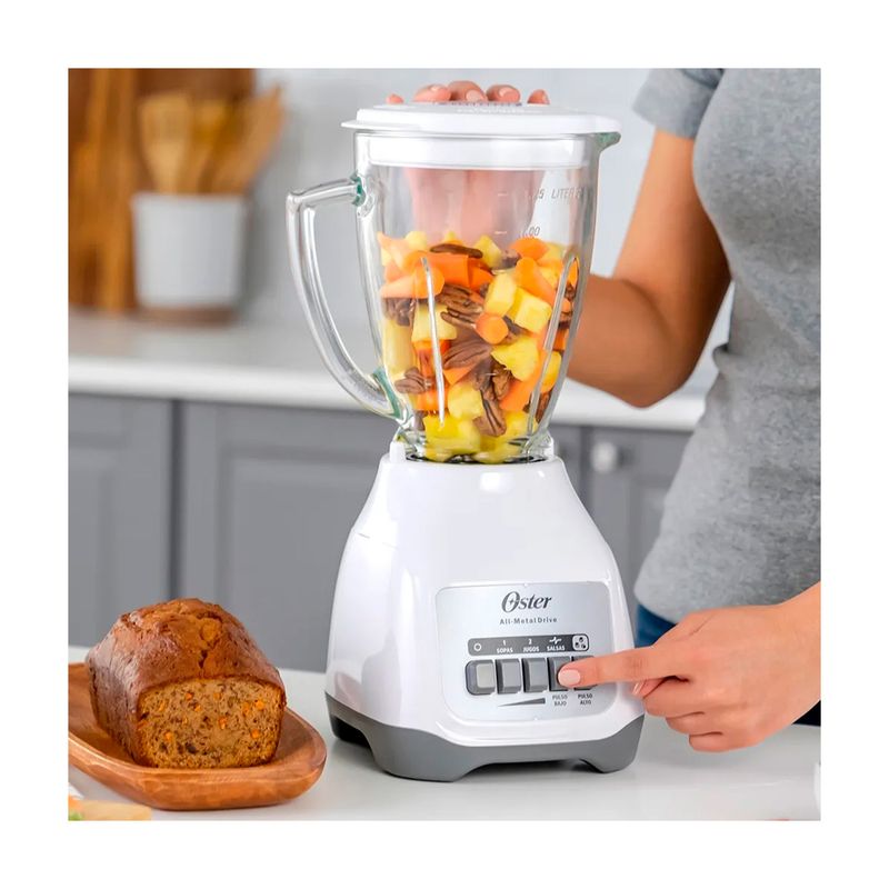 Licuadora vaso vidrio 1.5 L 800 W 2 vel blanca Oster, en uso con frutas picadas y mano operando los botones