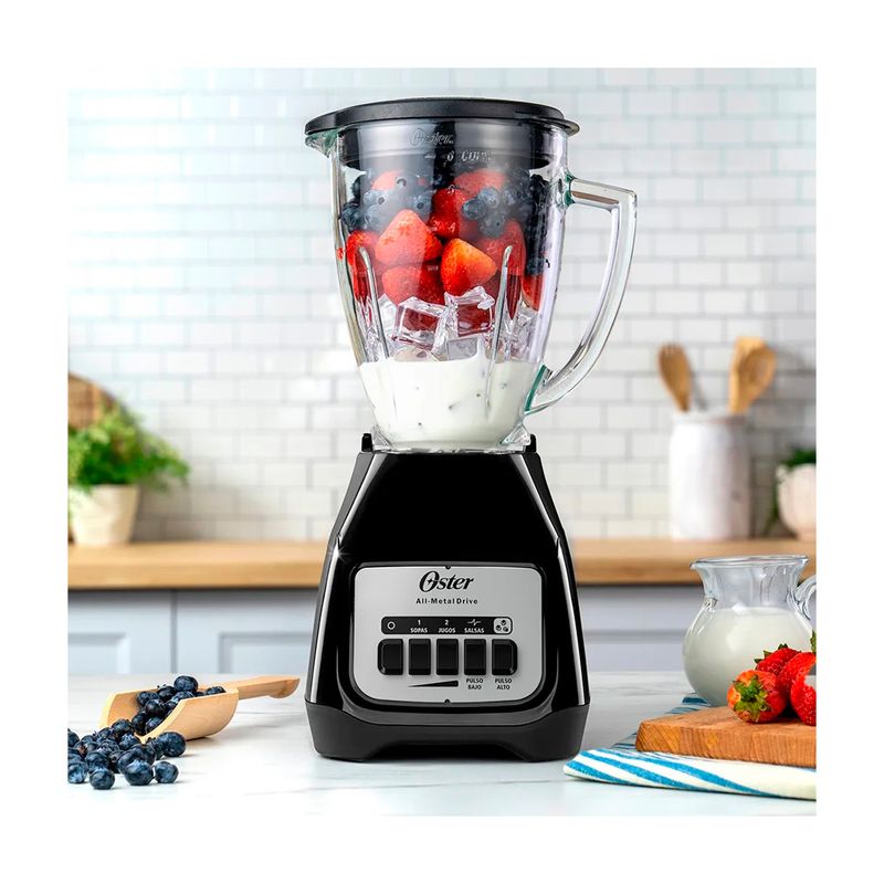 Licuadora vaso vidrio 1.5 L 800 W 2 vel negra Oster, en uso con frutas, hielo y leche sobre una mesa de cocina
