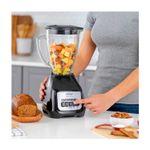 Licuadora vaso vidrio 1.5 L 800 W 2 vel negra Oster, usada por una persona con trozos de fruta dentro del vaso