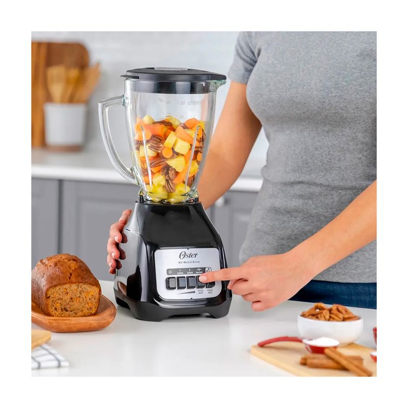 Licuadora vaso vidrio 1.5 L 800 W 2 vel negra Oster, usada por una persona con trozos de fruta dentro del vaso
