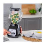 Licuadora vaso vidrio 1.5 L 800 W 2 vel negra Oster, en uso con brócoli y piña en el vaso sobre una encimera