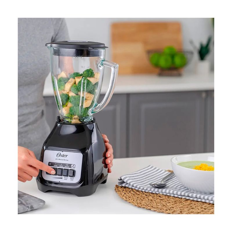 Licuadora vaso vidrio 1.5 L 800 W 2 vel negra Oster, en uso con brócoli y piña en el vaso sobre una encimera