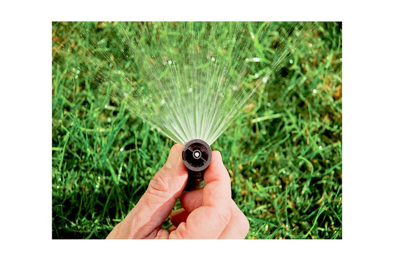 Boquilla para aspersor 15 pies ajustable 360 grados Rain Bird, sostenida por una mano mientras rocía agua sobre el césped
