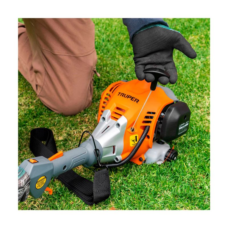 Desbrozadora gas 30 cc 17 pulgadas curva Truper, detalle del motor naranja sobre el césped con mano accionando el arranque
