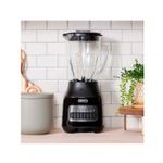 Licuadora vaso vidrio 1.5 L 800 W 6 vel gris Oster, vista frontal sobre encimera junto a una planta y recipiente